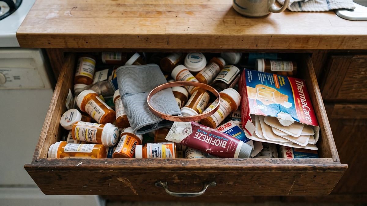 A messy drawer pulled open on a kitchen counter, full of pill bottles, a braided knee sleeve, and a thin shiny copper co